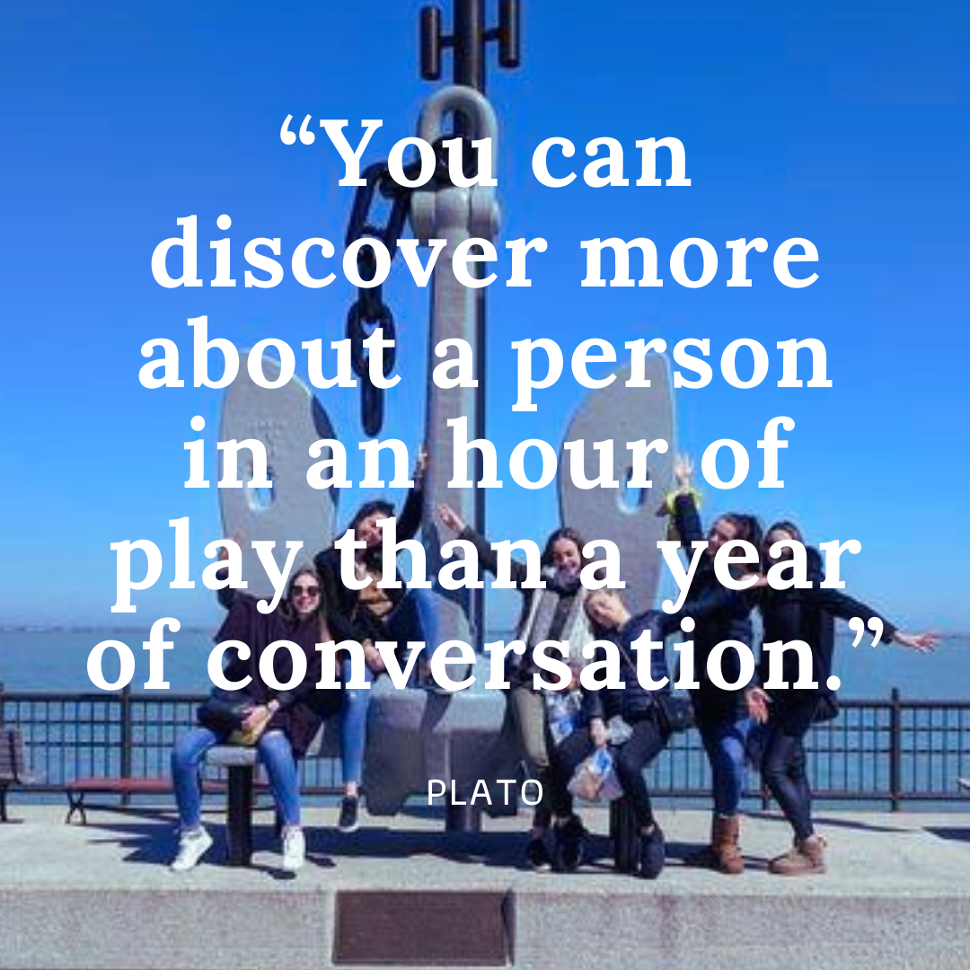 A group of 6 young people smiling and making fun poses in front of a giant ship anchor at least 12 feet tall and 10 feet wide, the location is a concrete sidewalk on a public pier with a low iron fence and a large lake in the background. Text over image reads "You can discover more about a person in an hour of play than a year of conversation. - Plato"